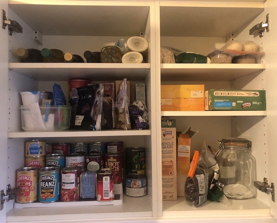 White kitchen cupboard with multiple shelves of tinned food, bottles and packets