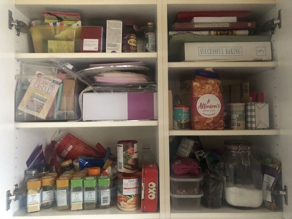 White kitchen cupboard with multiple shelves full of spice, baking ingredients, recipe books and assorted boxes