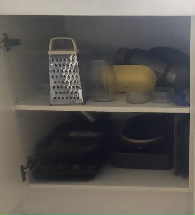 White kitchen cupboard with two shelves of baking tins, plastic storage boxs and a grater