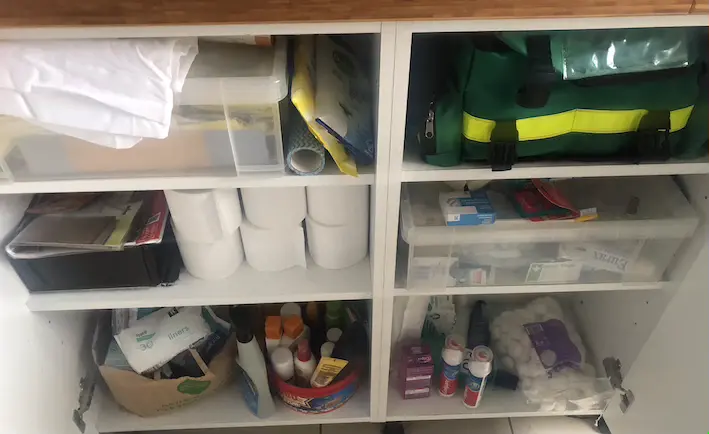 White kitchen cupboard with multiple shelves full of plastic boxes, first aid kits, toiletries and toilet rolls