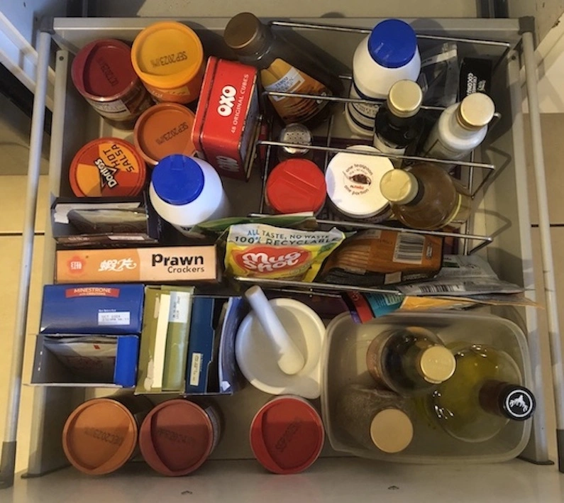 View from above of a white kitchen drawer, full of food jars, bottles and packets