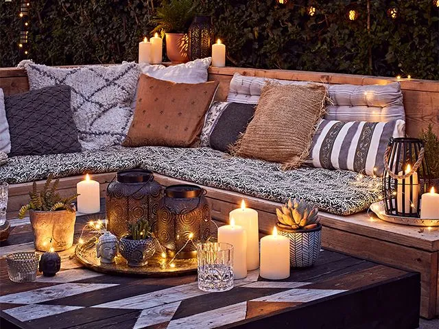 A cluster of lit candles and ornaments on a table in front of a corner seat covered in cushions