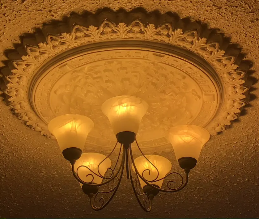 Illuminated 5 armed black metal chandelier with upturned glass globes, lit with an orange-toned light, below a patterned plaster ceiling rose.