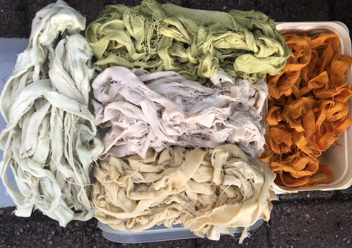 five piles of dyed cloth. Pale green to the left and orange to the right. Three piles in the centre range from pale green, through cream to light brown.