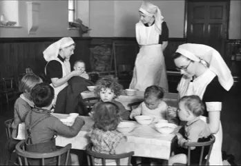 Black and white image of children eating at a table attended by nurses in uniforms and veils