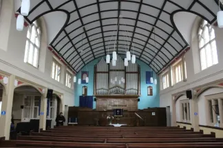 Church interior, mostly painted in shades of cream, showing pews in front of a raised wooden platform and organ pipes on a turquoise wall