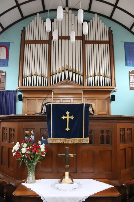 Organ pipes on a turquoise wall,above a wooden pulpit and table displaying a brass cross and vase of flowers