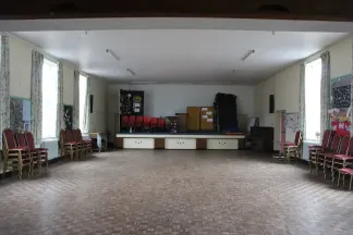 Interior of church hall, with wooden floor, large windows and stacked chairs on both sides, and a raised platform at the end