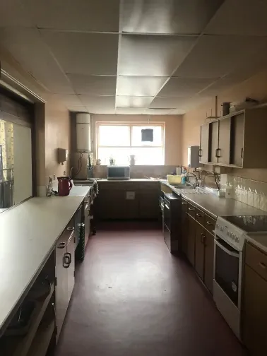 Galley kitchen in a church hall, a window at the end, a serving hatch on the left, cupboards and appliances on both sides