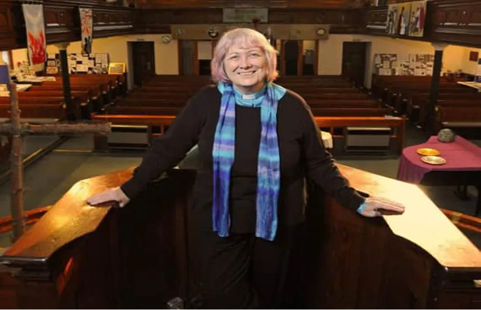 A female minister with blonde hair, wearing black and a purple scarf, standing in a pulpit facing away from the pews in a church