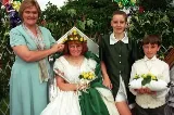 Girl in a white dress and dark green cloak, with yellow roses in her hair, sitting on a throne accompanied by a woman and two page boys