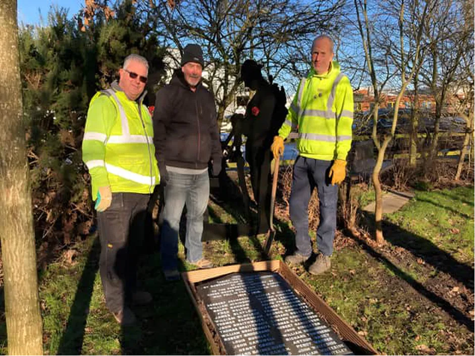 Three men in work clothes, standing beside a granite tablet inscribed with names, lying flat on grass beside some trees