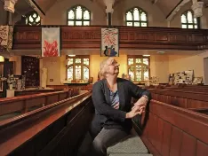 A woman sitting sideways in a pew of a traditional church, looking up towards the rear balcony