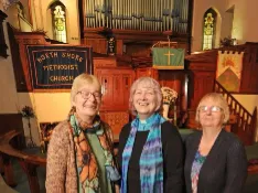 Three women standing in the upper balcony of a traditional church, organ pipes and pulpit visible behind them