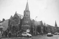 Black and white view of a church with a spire, people and cars passing by