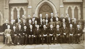 Monochrome picture of a group of 38 men, most wearing black suits and bow ties, some sitting, some standing, outside a church door