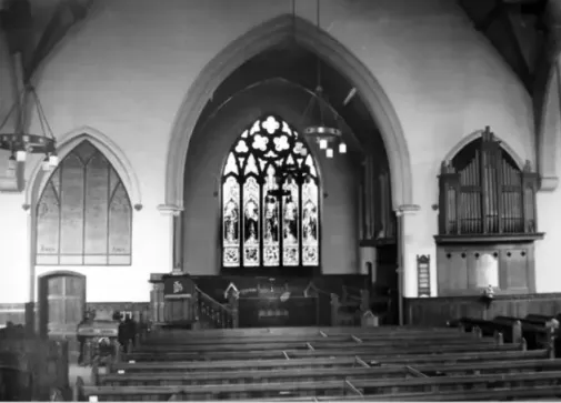 Monochrome interior of a church with pews in the foreground, a central stained glass window, organ pipes to the right and arched memorial to the left